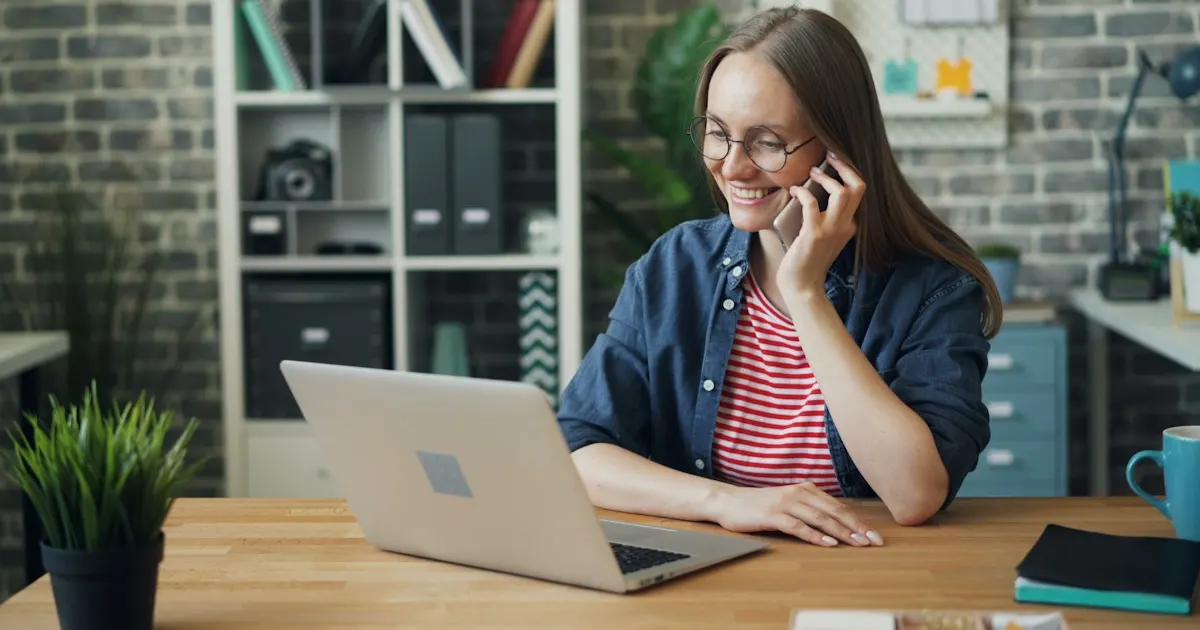 A smiling woman talking on her phone while looking at a laptop