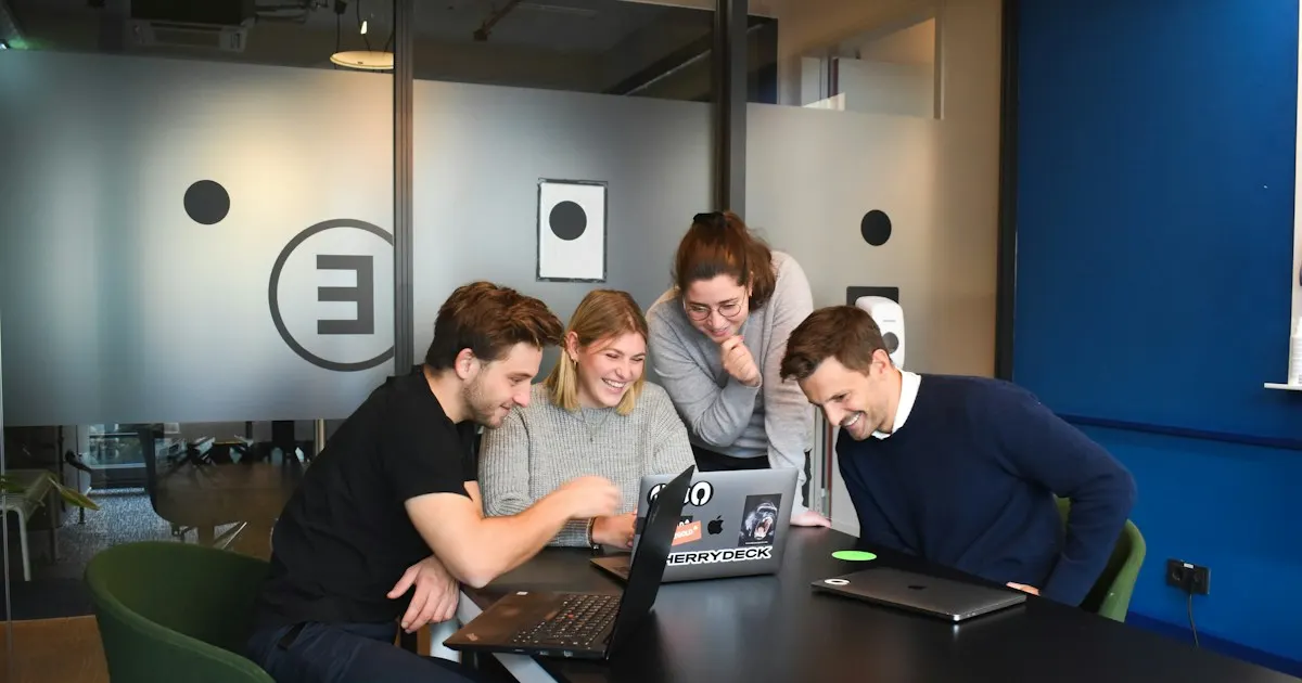 Four coworkers gathered around a laptop, smiling at something on the screen